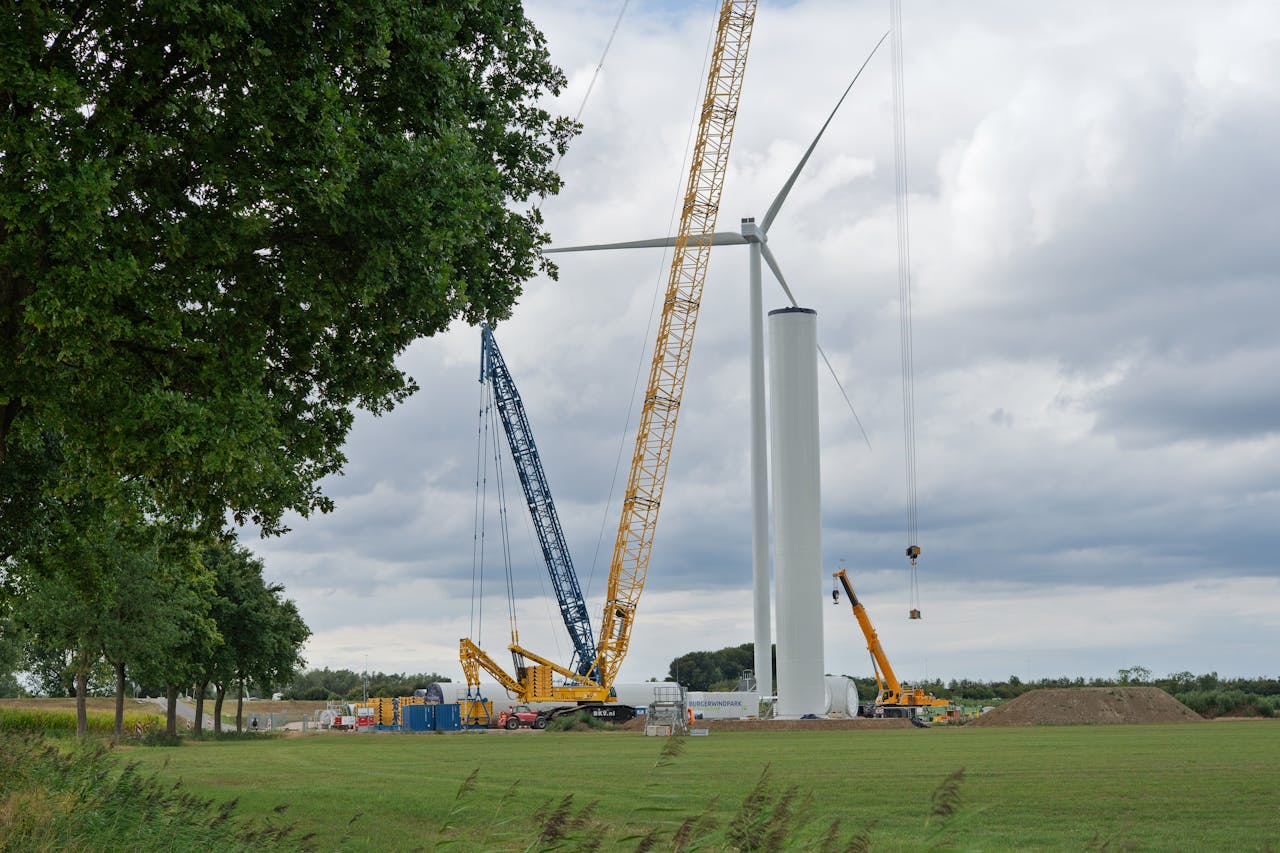 gallery-03 Cranes assembling a wind turbine in a grassy field under a cloudy sky.