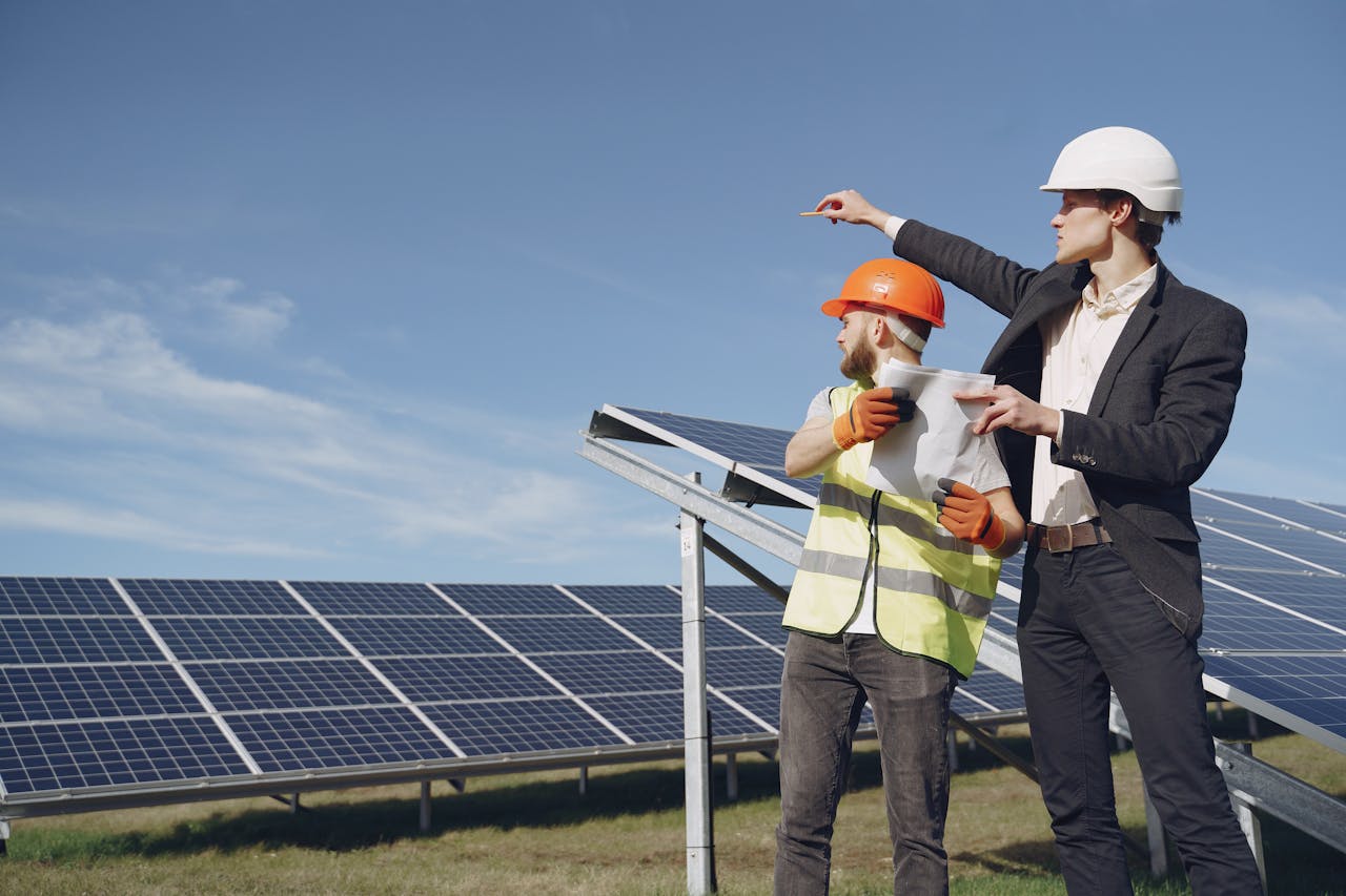 gallery-02 Young businessman in formal outfit and foreman in yellow vest looking away while discussing working process against solar panels